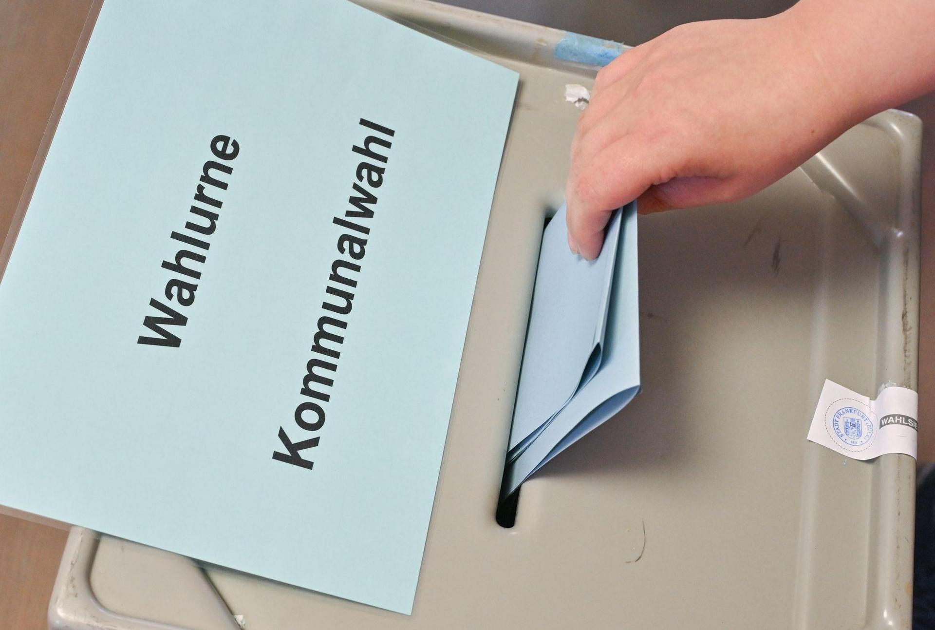 Eine Hand steckt einen Stimmzettel in eine Wahlurne mit der Aufschrift „Wahlurne Kommunalwahl“. (Symbolbild: Patrick Pleul/dpa)